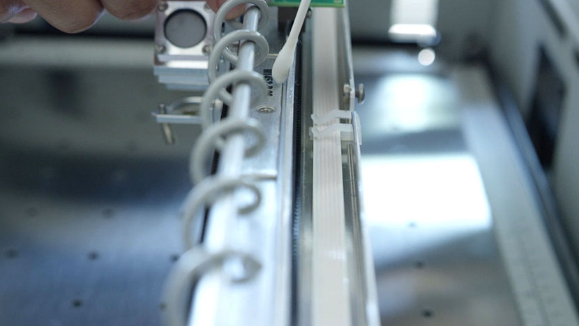 Run the cotton swab along the front of the encoder strip. Cleaning the front of the linear encoder strip.